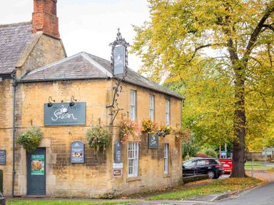 Exterior of a stone building with signs and hanging flower baskets next to a tree at The Swan in Moreton in Marsh