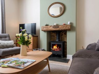 A living room with a wood burning stove under a wooden beam mantel a round mirror above and a television on a wooden cabinet in Wall Cottage in Burford