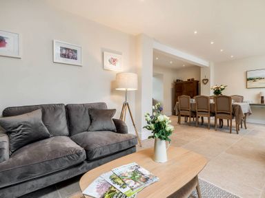 A living room with a gray sofa a wooden coffee table with flowers and magazines and a dining area with a table and six chairs at Wall Cottage in Burford