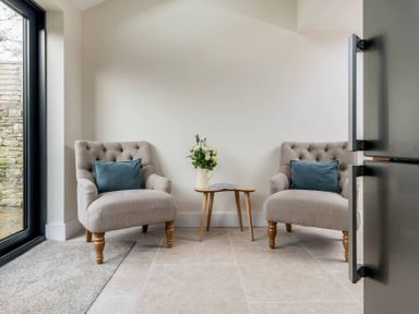 Two beige armchairs with blue cushions and a small wooden table with flowers and a magazine in a room with tiled floor at Wall Cottage in Burford