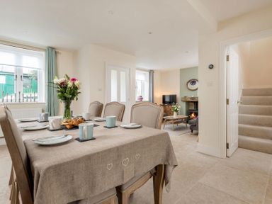 A dining area with a table set with cups and plates and a vase of flowers next to a living room with a fireplace and TV at Wall Cottage in Burford