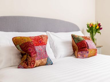 A bed with white sheets and two colorful patchwork pillows with a vase of flowers on a bedside table at Wall Cottage in Burford