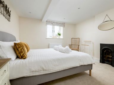 A bedroom with a bed with white linens and brown pillows two rolled towels on the bed a wooden chair a window with floral curtains and a fireplace at Wall Cottage in Burford