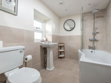 A bathroom with a toilet sink corner shelf bathtub with shower and round mirror at Wall Cottage in Burford