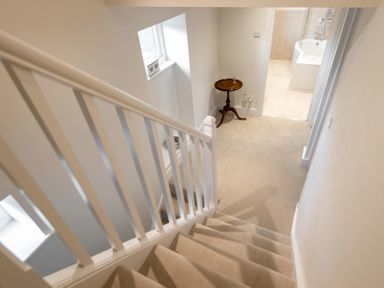 A stairway with beige carpet leading down to a small landing with a wooden round table next to a window and an open doorway to a bathroom at Wall Cottage in Burford