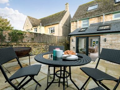 A patio area with black woven chairs and table set with croissants and mugs outside a stone house with glass doors at Wall Cottage in Burford