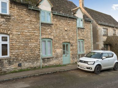 A stone house with green doors and windows and a white car parked outside on the street at Wall Cottage in Burford