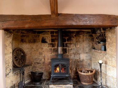 A stone fireplace with a wood burning stove a basket of firewood and fireplace tools at Little Gables in Broadway