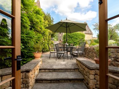 A stone patio with a table and chairs under an umbrella surrounded by plants and stone walls at Little Gables in Broadway