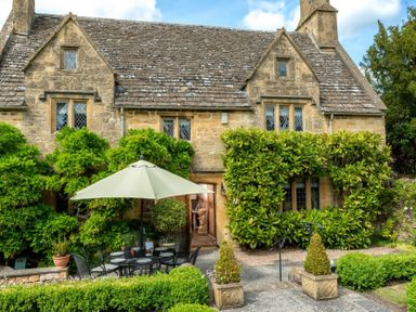An outdoor seating area with a table umbrella and chairs in front of a stone house with windows and greenery at Little Gables in Broadway