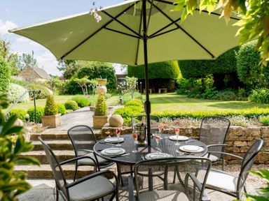 An outdoor patio dining area with a table, chairs, plates, glasses, and an umbrella in a garden at Little Gables in Broadway