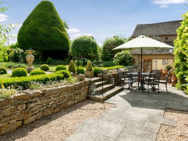A garden patio with an outdoor dining set and umbrella next to a stone wall and trimmed bushes at Little Gables in Broadway