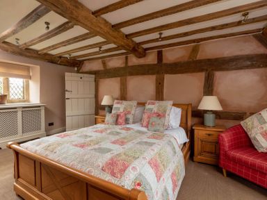A bedroom with a wooden bed and patchwork bedding two nightstands with lamps a red armchair exposed wooden beams on the ceiling and walls at Little Gables in Broadway