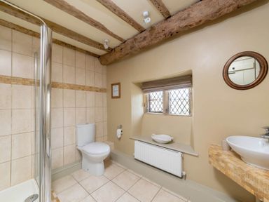 A bathroom with a toilet a shower a sink a window and wooden ceiling beams at Little Gables in Broadway