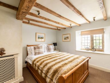 A bedroom with a wooden bed with plaid pillows and a fur blanket wooden ceiling beams and a window with a model bicycle at Little Gables in Broadway