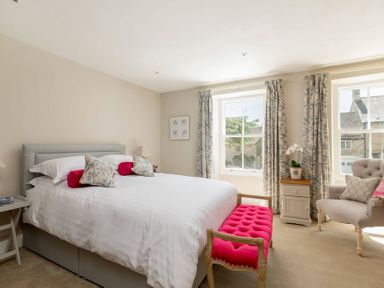 A bedroom with a large bed with white bedding and red cushions a pink bench at the foot of the bed floral curtains two windows an armchair and a small bedside cabinet with a plant Copperbeech Stow-on-the-Wold