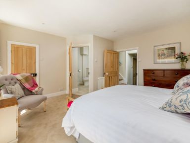 A bedroom with a bed pillows a wooden dresser an armchair with a blanket flowers and doors at Copperbeech in Stow-on-the-Wold