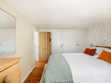 A bedroom with a bed covered with white bedding and orange pillows a wooden dresser a wall mirror wooden door and white wardrobes at Copperbeech in Stow on the Wold