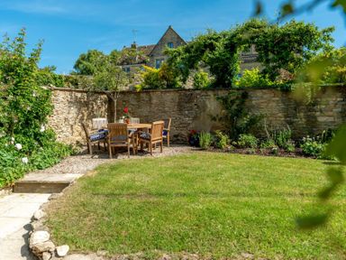 A garden with a wooden dining table set on gravel beside a stone wall covered with plants at Copperbeech in Stow-on-the-Wold