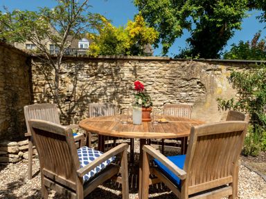 An outdoor seating area with a round wooden table and six wooden chairs with cushions surrounded by stone walls and trees at Copperbeech in Stow-on-the-Wold