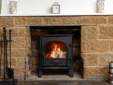 A lit wood burning stove in a stone fireplace with a clock on the mantel at Holly Cottage Middle Tysoe in Tysoe