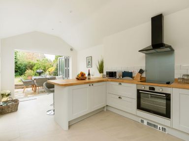 A kitchen with white cabinets wooden countertops an oven microwave and an open door leading to a garden with a patio table and chairs at Holly Cottage Middle Tysoe in Tysoe