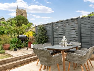 An outdoor patio with a table and four wicker chairs near a barbecue grill and garden with a church tower visible in the background at Holly Cottage Middle Tysoe in Tysoe