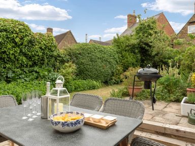 A garden patio with a table set with glasses bowl and lantern and a barbecue grill and dense bushes and houses in the background at Holly Cottage Middle Tysoe in Tysoe