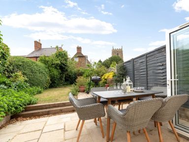 An outdoor patio with a dining table and six wicker chairs next to a garden and black fence at Holly Cottage Middle Tysoe
