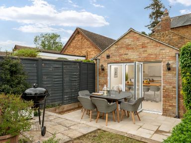 A backyard patio with a dining table and chairs next to a brick house with open glass doors at Holly Cottage Middle Tysoe in Tysoe