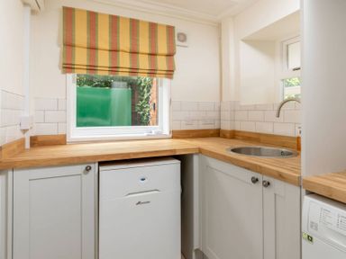 A kitchen with wooden countertops white cabinets a small white fridge a round stainless steel sink and windows with a striped roman blind at Holly Cottage Middle Tysoe in Tysoe