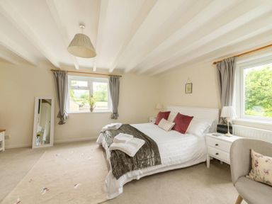A bedroom with a double bed two windows with curtains bedside tables a standing mirror and an armchair at Holly Cottage Middle Tysoe in Tysoe
