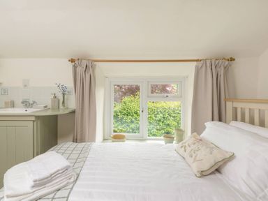 A bedroom with a bed, floral pillow, folded towels, window with curtains, and a small sink area at Holly Cottage Middle Tysoe in Tysoe