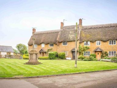 A thatched roof cottage with multiple doors and windows surrounded by a lawn with a stone cross monument and a wooden utility pole at Holly Cottage Middle Tysoe in Tysoe