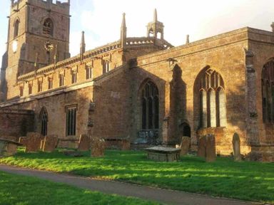 An old stone church with a clock tower and arched windows surrounded by gravestones and green grass at Holly Cottage Middle Tysoe in Tysoe