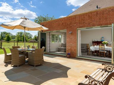 A patio with a table and chairs under an umbrella next to a brick house with open doors showing a dining area and living room at Mount View in Childswickham