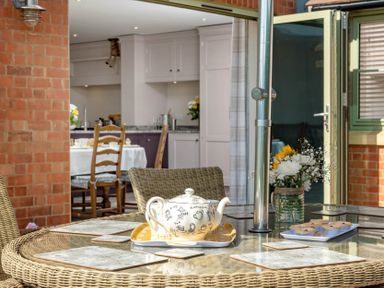 An outdoor glass table with a teapot cupcakes and flowers with a view into a kitchen with a dining table and chairs at Mount View in Childswickham