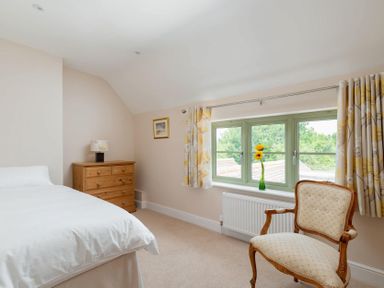 A bedroom with a bed white bedding wooden dresser lamp floral curtains armchair and a window with a vase and flowers at Mount View in Childswickham