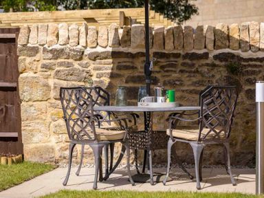An outdoor patio with a metal table and four metal chairs with cushions against a stone wall at Millbank Cottage in Mickleton