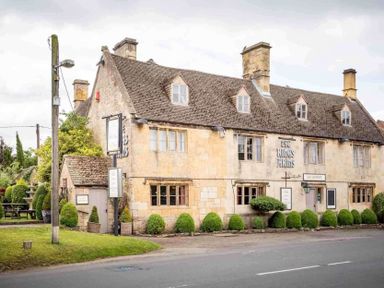 An old stone building with multiple windows and chimneys along a roadside with bushes in front at Millbank Cottage in Mickleton