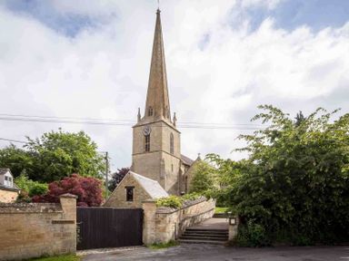 A stone church with a tall clock tower and steeple surrounded by greenery and stone walls at Millbank Cottage in Mickleton