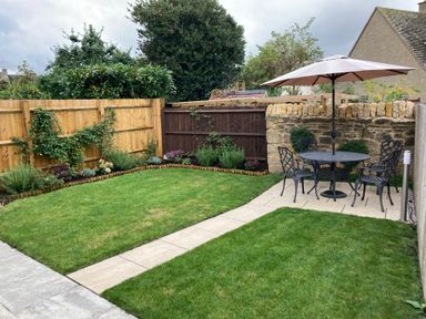 A garden with a lawn, flower beds along wooden and stone fences, a patio with a table, four chairs, and a large umbrella at Millbank Cottage in Mickleton