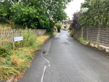 A narrow paved road lined with wooden fences and trees with houses visible at the end of the road at Millbank Cottage in Mickleton