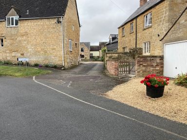 A street view of stone houses with a gravel area and a flower pot with red flowers at Millbank Cottage in Mickleton