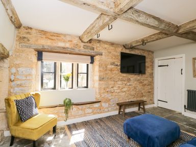 A room with stone walls a yellow chair a blue ottoman a wall-mounted tv and wooden ceiling beams at Millbank Cottage in Mickleton