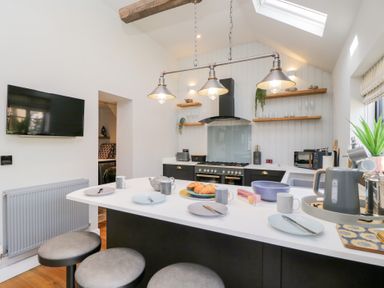 A kitchen with a white island counter set with plates mugs and croissants a black stove and hood wooden shelves and a skylight at Millbank Cottage in Mickleton