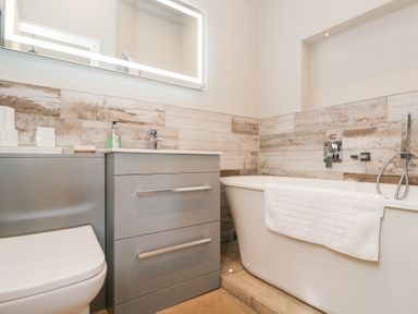 A bathroom with a bathtub, a towel hanging on it, a sink with a cabinet below, a wall mirror, and a toilet at Millbank Cottage in Mickleton