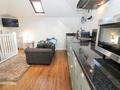 A kitchen with green cabinets a countertop and a microwave next to a living area with a gray sofa a side table and a wall mounted TV at The Bath House in Blockley