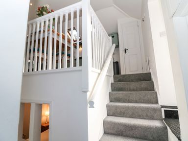 A staircase with gray carpet and white railing leading up to a white door with a glimpse of a dining area through the railing at The Bath House in Blockley