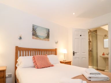 A bedroom with a wooden bed frame, white bedding, a pink cushion, bedside tables, a lamp, and an open door showing an ensuite bathroom at The Bath House in Blockley